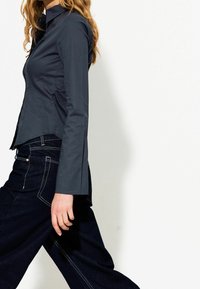 Eine Frau geht in einem engen, dunkelblauen Langarmshirt und lockeren, dunklen Jeans mit weißen Nähten vor einem weißen Hintergrund.