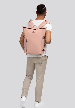 Man in een wit t-shirt en beige broek, die een grote roze rugzak met meerdere ritsen en banden draagt, staande tegen een grijze achtergrond.
