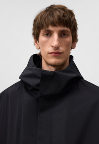 Svart vattentät jacka med hög krage, minimalistisk design, slät textur och dolda dragkedjor. Ljus reflekteras på tygets yta.