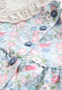 Blau gemusteter Stoff mit Blumenmuster in Hellblau, Akzenten in Pink und Grün, mit einem weißen Spitzenkragen und blauen Knöpfen. Weiches, strukturiertes Material.