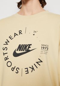 Beige sweatshirt med en stor svart Nike-logga, cirkulärt textdesign och riktningselement med "Sedan 1972" tryckt på framsidan.