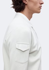 Homme de profil portant une chemise blanche avec une poche à rabat boutonnée sur la partie supérieure de la manche, sur un fond uni clair.