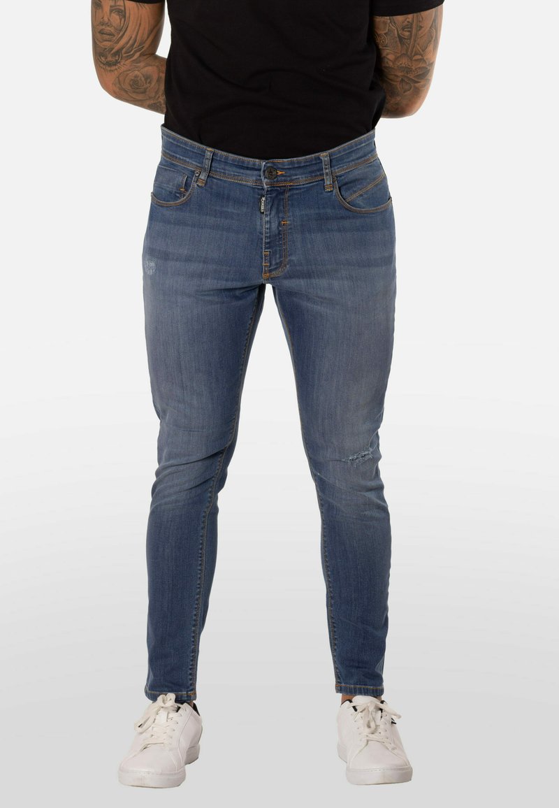 Jeans de mezclilla azul con corte ajustado, que presentan bolsillos delanteros, desgaste ligero en el muslo y un diseño clásico de cinco bolsillos, combinados con zapatillas blancas.