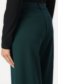 Hoge taille donkergroene broeken met een zachte textuur, voorzien van twee achterzakken en een getailleerd ontwerp, gedeeltelijk zichtbaar met een zwart topje.