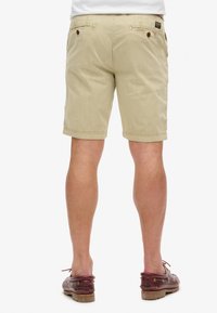 Pantaloni corti beige in cotone con taglio dritto, dotati di due tasche posteriori con chiusura a bottone e una texture sottile. Indossati con scarpe da barca marroni.