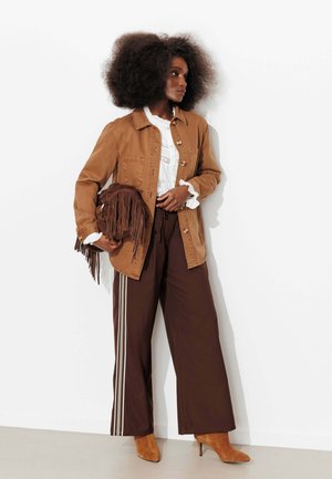 Femme aux cheveux bouclés volumineux portant une veste marron sur un chemisier blanc, un pantalon large marron avec des bandes latérales, des bottes couleur sable, et tenant un sac à franges.