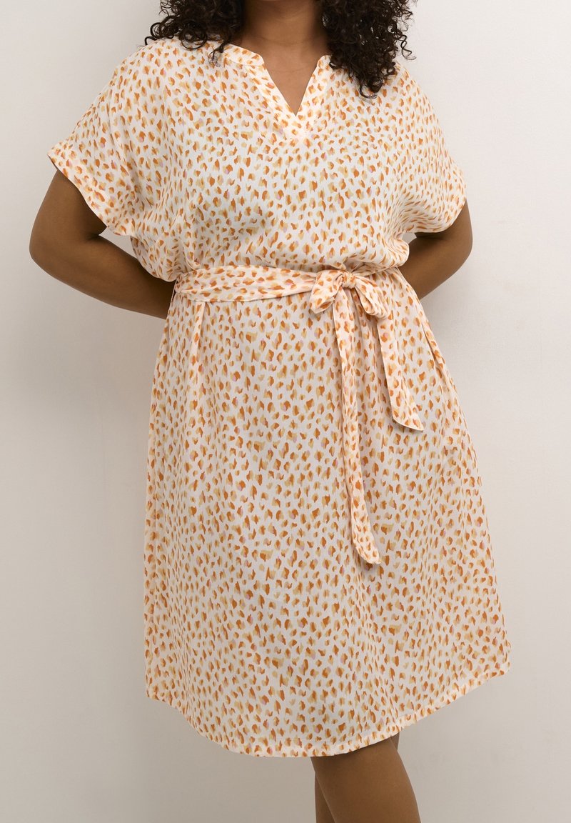 Femme portant une robe blanche avec des taches abstraites orange et une ceinture nouée à la taille, debout devant un fond clair uni.
