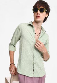 Camisa abotonada verde claro con un patrón geométrico texturizado, mangas remangadas y un collar. Lleva puestas gafas de sol y un collar.