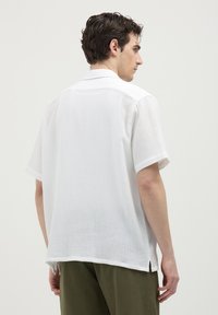 Giovane uomo con capelli castani ricci che indossa una camicia bianca a maniche corte di tessuto testurizzato e pantaloni verde oliva, rivolto di lato su uno sfondo neutro.
