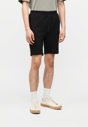 Mann trägt schwarze Shorts mit Kordelzug, weiße Socken und weiße Turnschuhe und steht vor einem schlichten hellen Hintergrund.