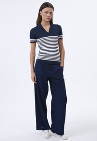 Polo shirt lavorato a maglia di colore navy con motivo a strisce bianche e rosse, maniche corte e scollatura a V, abbinato a pantaloni larghi navy e sneakers bianche.