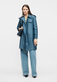 Γυναίκα που φοράει παλτό μπλε με σχέδια, ανοιχτόχρωμα μεγάλα παντελόνια σε μπλε απόχρωση, μαύρα παπούτσια και κρατάει μια μαύρη τσάντα στον ώμο.