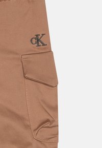 Pantalon cargo Calvin Klein en tissu beige clair avec une poche latérale à rabat, des détails de couture subtils et un logo brodé en noir.