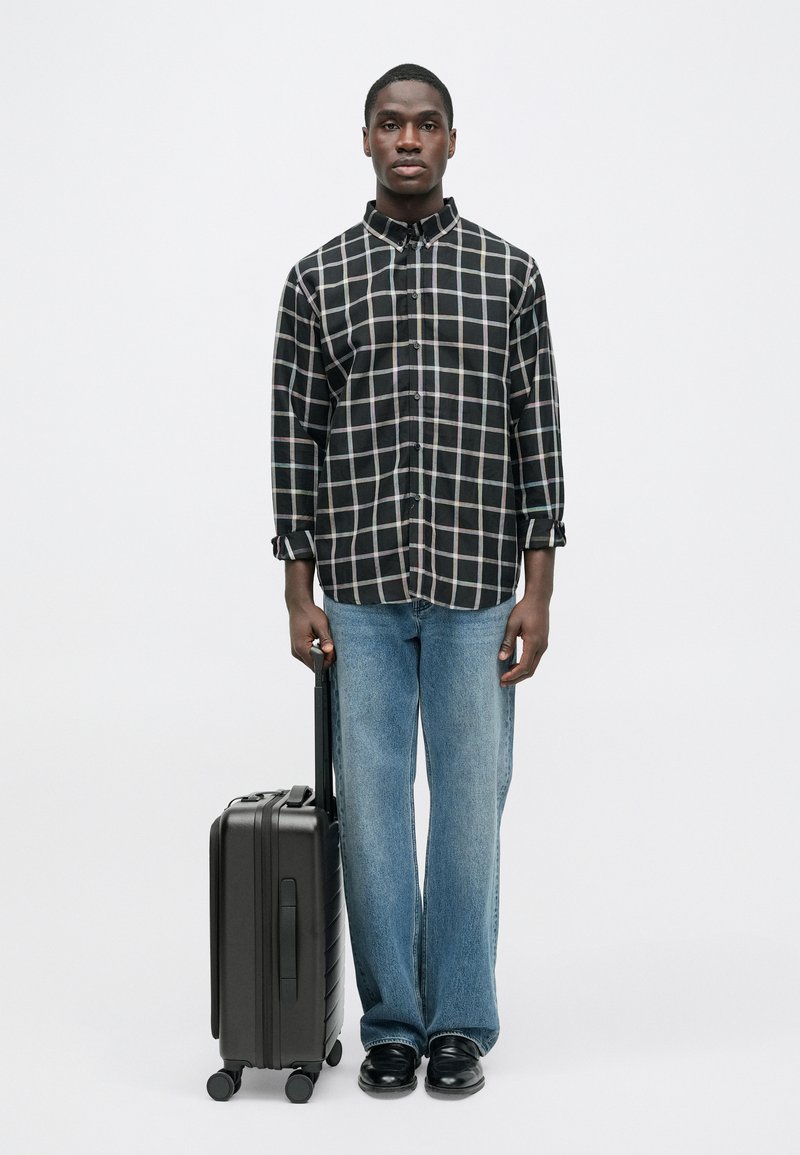 Black and white checkered button-up shirt, light blue jeans, black shoes, holding a compact black suitcase with a retractable handle.