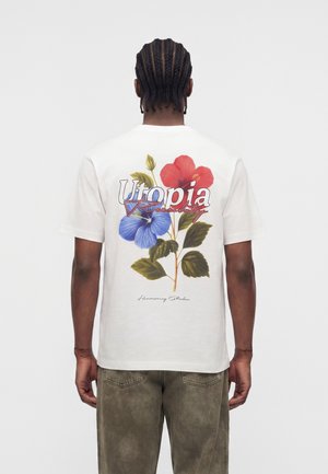Mann med fletter iført hvit T-skjorte med rød og blå blomstergrafikk og teksten "Utopia" på baksiden, kombinert med falmede bukser.