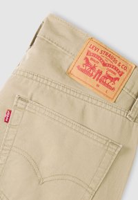 Μπεζ βαμβακερό παντελόνι με δερμάτινη ετικέτα που αναγράφει "Levi Strauss & Co." και κόκκινη ταμπέλα στην τσέπη· διαθέτει χαρακτηριστική καμπυλωτή ραφή.