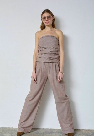 Žena nosiaca bezový top bez ramienok s rebrovanou štruktúrou a široké nohavice, stojaca pri bielej stene, doplnená slnečnými okuliarmi, zlatými šperkami a hnedými topánkami.