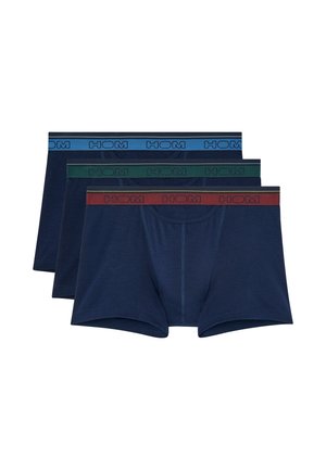 Hombre musculoso de pie descalzo con bóxers azul marino con cinturilla roja sobre un fondo liso gris claro.