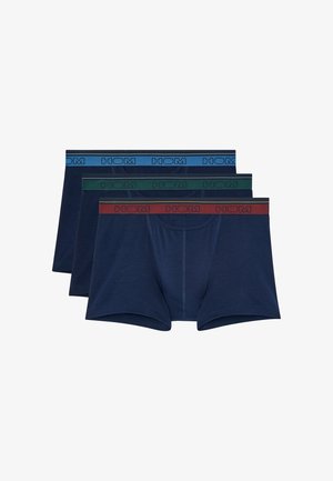 Hombre musculoso de pie descalzo con bóxers azul marino con cinturilla roja sobre un fondo liso gris claro.