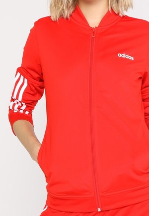 Frau trägt eine rote Adidas Jacke mit Reißverschluss, weißen Streifen an den Ärmeln und Adidas-Logo auf der Brust, Hand in der Tasche, blickt nach vorne.