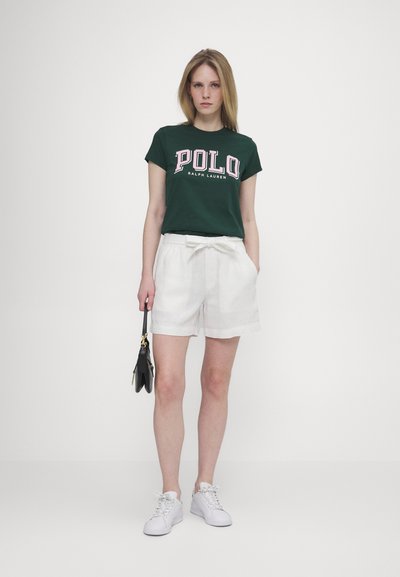 Grønn t-skjorte med kort erm og "POLO" grafikk, kombinert med hvite linshorts. Modellen holder en svart håndveske og har på seg hvite joggesko.