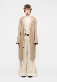 Άτομο στέκεται στραμμένο μπροστά, φορώντας μια μπεζ μακριά καμπαρντίνα πάνω από ένα λευκό τοπ, φαρδιά κρεμ παντελόνια, καφέ ζώνη και μαύρα παπούτσια, με φόντο απλό.