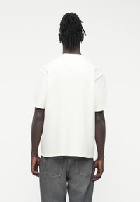 Camiseta blanca de algodón con mangas cortas, corte holgado y escote redondeado. Se lleva con unos vaqueros grises de mezclilla, que tienen bolsillos traseros.