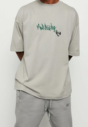 T-shirt beige oversize avec texte vert "Multiply" et petit graphique d'une main tenant une bombe de peinture sur la poitrine, porté avec un pantalon beige à cordon.
