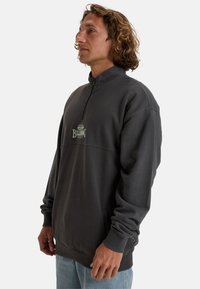 Pullover en polaire gris foncé avec un col haut et une fermeture éclair quart. Présente un logo brodé sur le devant, des poignets côtelés et des poches latérales.