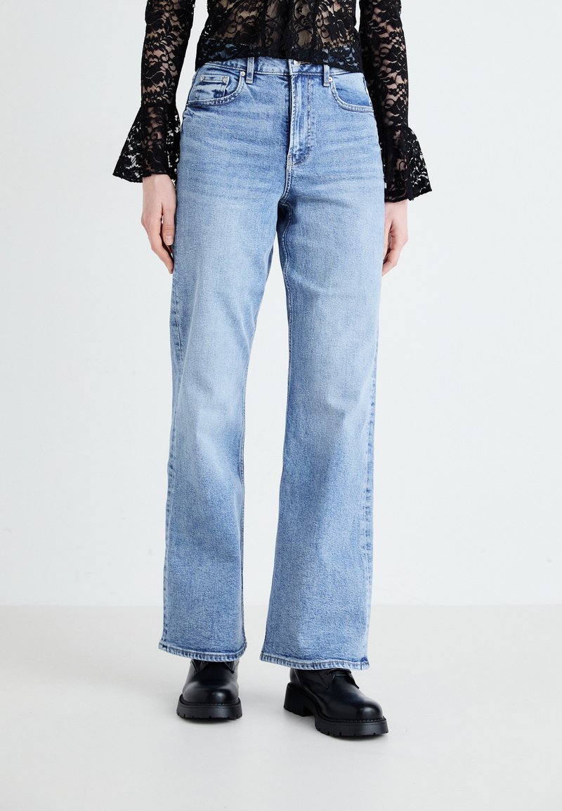 Jean évasé bleu clair en denim avec une finition décolorée, doté de cinq poches et d'une fermeture éclair, associé à des bottes à plateforme noires.