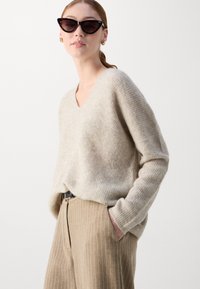 Light beige, v-neck sweater made of soft knit material, paired with beige pinstriped trousers. Model poses with hands in pockets, wearing dark sunglasses.