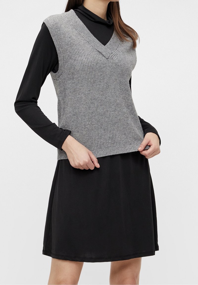 Femme portant un gilet gris en maille sans manches à col en V par-dessus une robe noire à manches longues, les mains touchant l'ourlet du gilet.