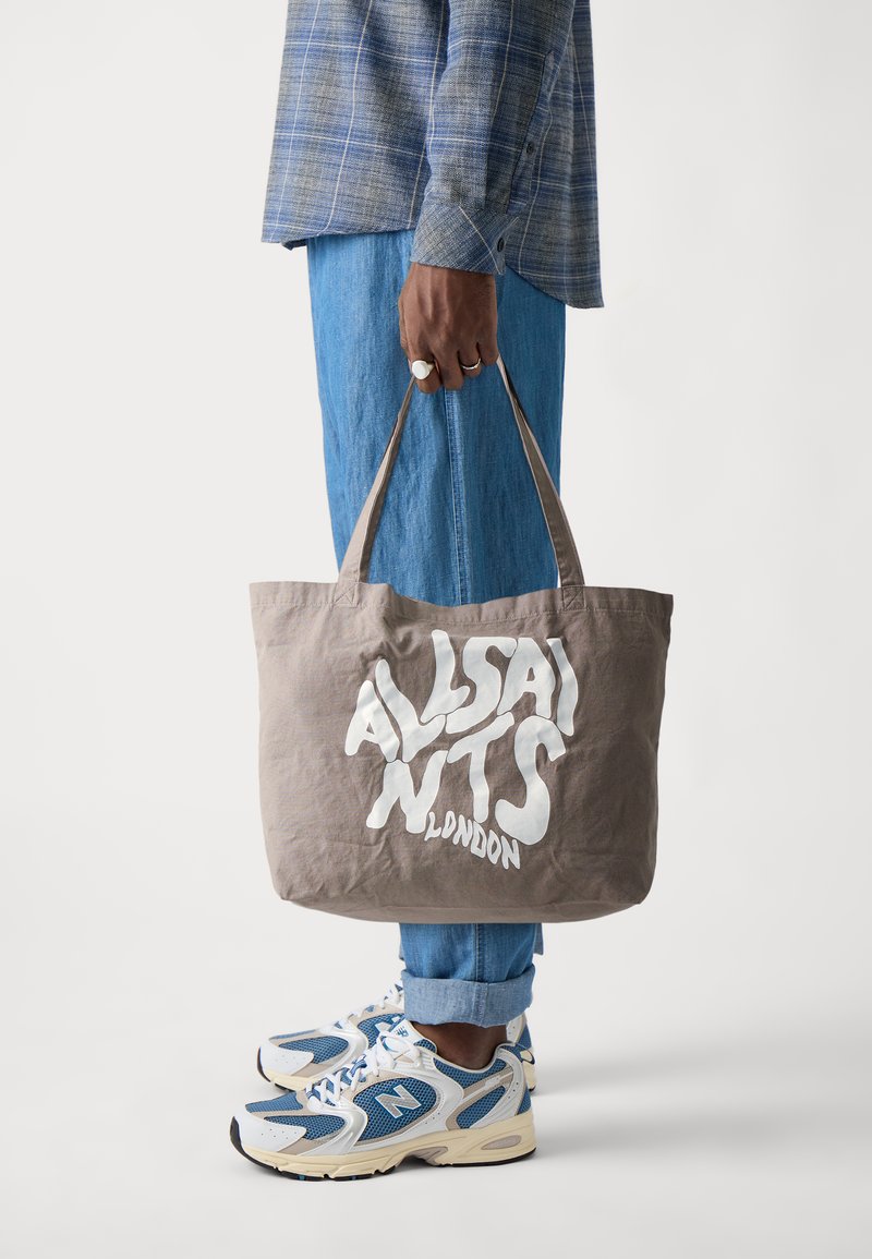 Grau farbener Stoffbeutel mit weißem "ALLSAINTS LONDON"-Text, strukturierte Oberfläche und langen Trägern, gehalten von einer Person, die blaue Jeans und Sneaker trägt.