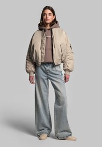 Beige bomberjakke med ribbede manchetter, brun hættetrøje under, parret med lyseblå vidde jeans og beige sneakers. Ingen bemærkelsesværdige mønstre.