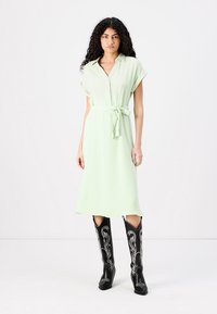 Rochie midi verde deschis cu un decolteu cu nasturi, mâneci scurte și un cordon legat în talie. Purtați cu cizme cowboy negre cu broderie albă.