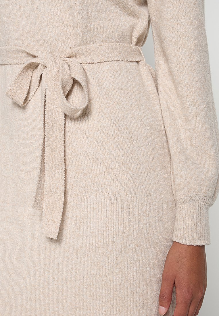 Robe en tricot beige, avec une texture douce, des manches longues et une ceinture nouée à la taille pour un ajustement réglable. Motif subtil chiné sur l'ensemble.