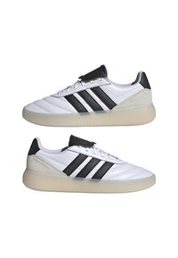 Chaussures de sport blanches avec des rayures noires, tige en cuir lisse, embout texturé et semelle en caoutchouc transparent, avec logo sur le talon.