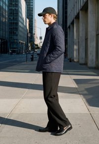 Jeune homme portant une casquette noire, une veste matelassée bleu marine, un pantalon noir et des mocassins, debout sur un trottoir en ville, les mains dans les poches.
