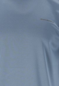 Vaaleansininen urheilupaita, joka on valmistettu teksturoidusta kankaasta; siinä on pieni tummanharmaa "ENDURANCE" -logo. Sujuvassa ja joustavassa muotoilussa on sopiva urheiluun.