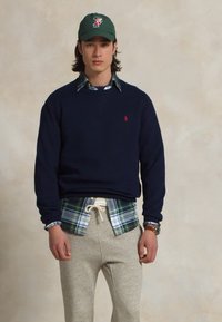 Tamsiai mėlynas džemperis su raudonu emblema, uždėtas ant žalsvai mėlyno languoto marškinėlių, derinamas su pilkais sportiniais džinsais ir žaliu kepurėle.