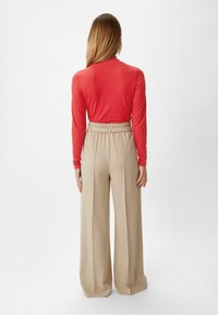 Vrouw met lang blond haar staat met haar rug naar de kijker gekeerd, gekleed in een koraalkleurige blouse met lange mouwen die in hoge beige wijde broek is gestoken.