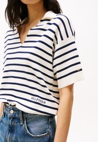 Ριγέ βαμβακερό polo μπλούζα σε λευκό και ναυτικό, με V-λαιμόκοψη, κοντά μανίκια και το "HILFIGER" κεντημένο μπροστά. Συνδυάζεται με τζιν σορτς.