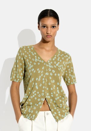 Młoda kobieta z rękami w kieszeniach, ubrana w zieloną krótką bluzkę z guzikami z jasnoniebieskim kwiatowym wzorem i białe spodnie.