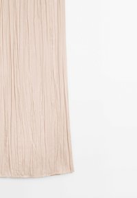 Tissu plissé beige clair suspendu verticalement avec des plis doux et une texture lisse visible sur un fond blanc.