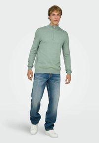 Lichtgroene trui met ritskraag gemaakt van zachte stof, gecombineerd met blauwe spijkerbroek en witte sneakers, voor een casual uitstraling.