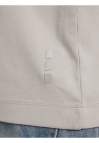T-shirt beige chiaro realizzato in tessuto morbido, con tre piccoli accenti rettangolari testurizzati sul lato sinistro vicino alla cucitura.