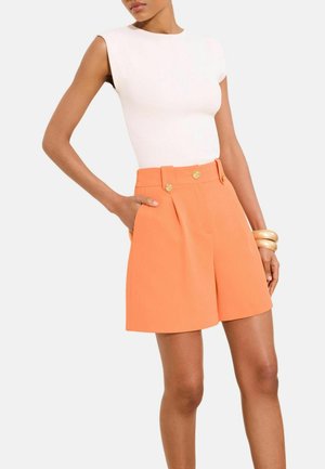 Frau trägt ärmelloses weißes Oberteil und hochtaillierte orangefarbene Shorts mit goldenen Knöpfen, dazu große goldene Armbänder am rechten Handgelenk.