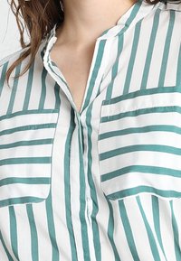 Camicia con bottoni caratterizzata da strisce verticali verde mare e bianche, con colletto e due tasche sul petto, realizzata in tessuto leggero.