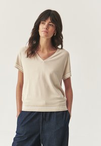 Beige rövidujjú, V-nyakú top, amely testhez álló szabású, textúrált pamut anyagból készült, és csíkos díszítéssel rendelkezik az ujjakon. Sötétkék nadrággal párosítva.
