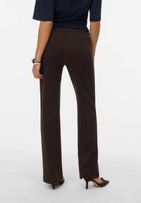 Brune, høytlivede bukser med rette ben laget av glatt stoff. Brukes med svart topp med korte ermer og brune sko med hæl, sett bakfra.